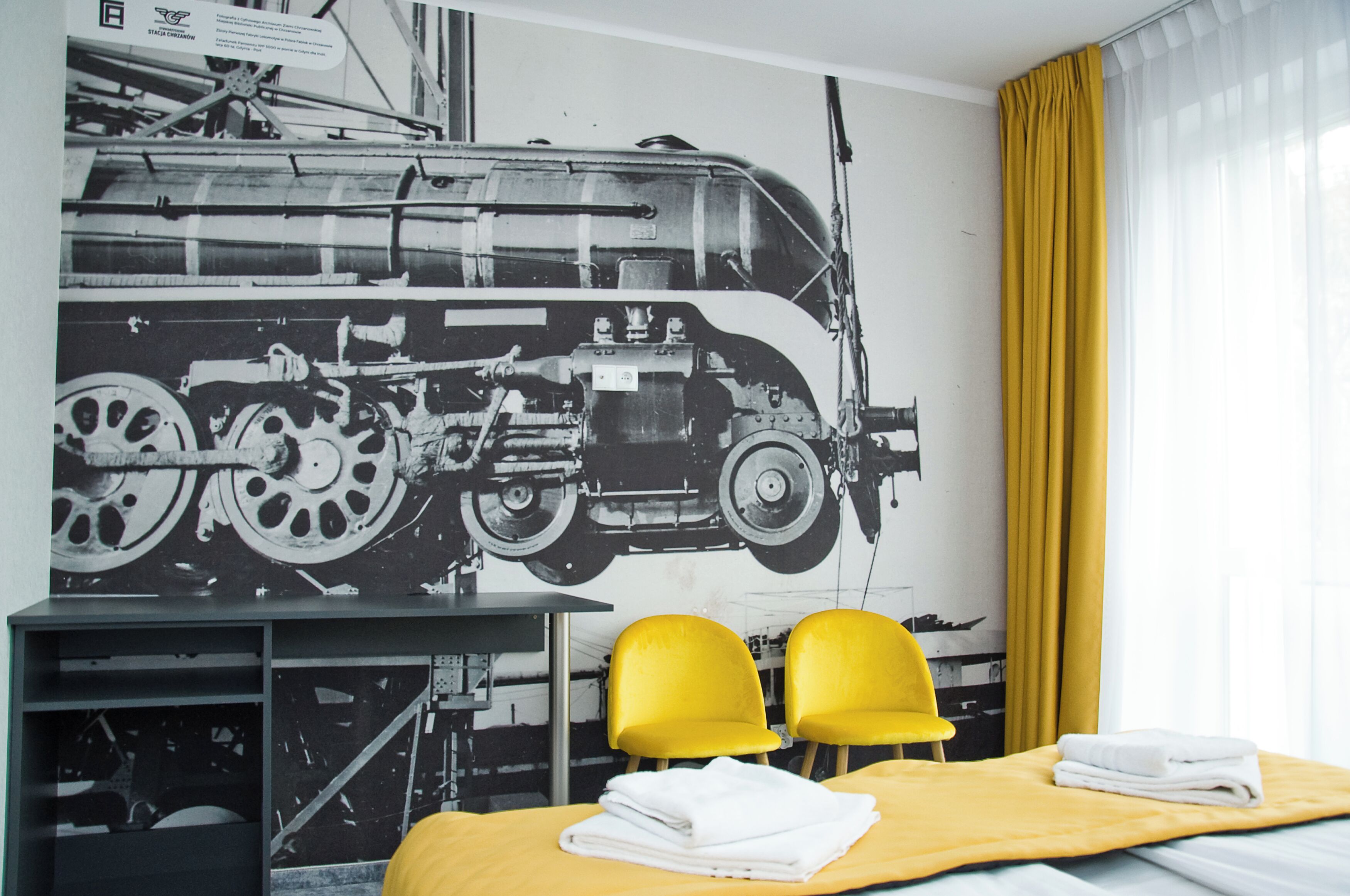 Une chambre d’hôtel avec un lit, deux chaises jaunes, un bureau et une grande fresque murale en noir et blanc représentant un train.