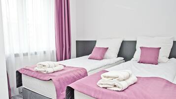 Dos camas individuales con ropa de cama blanca y morada, un cabecero gris y una ventana con cortinas de encaje.