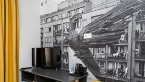 A modern room with a flat-screen TV, a small refrigerator, and two yellow chairs. A large mural of a building scene is on the wall. - ParkApartamenty (Chrzanow)