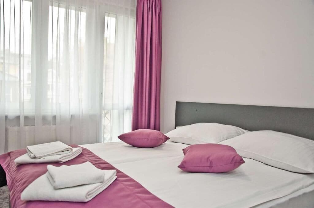 Ein ordentlich gemachtes Bett mit weißer und rosa Bettwäsche, ein grauer Kopfteil und ein Fenster mit durchscheinenden Vorhängen.