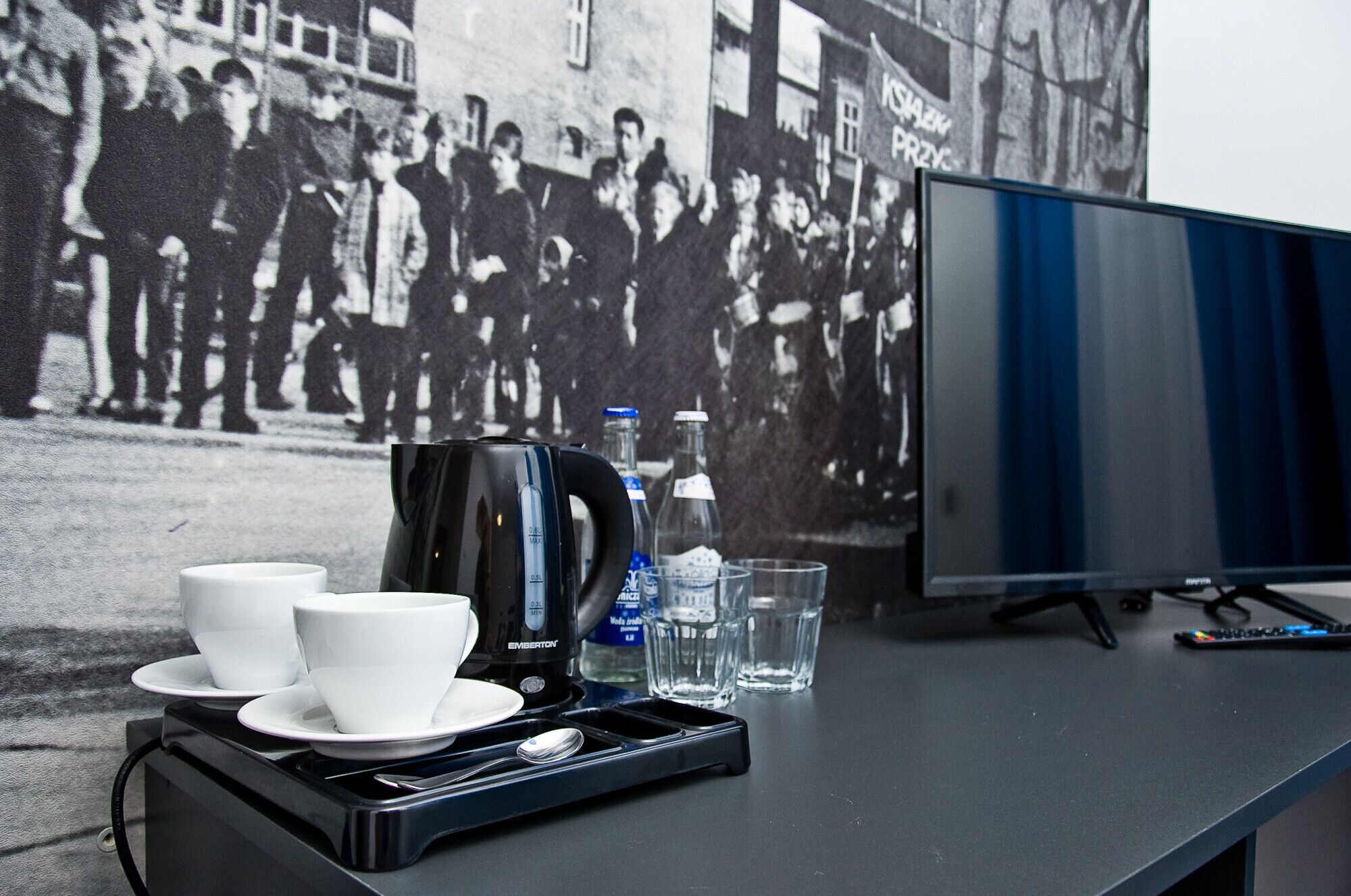 Een tafel met een koffiezetapparaat, twee kopjes, een dienblad en een glas water. Een televisie staat op de tafel.