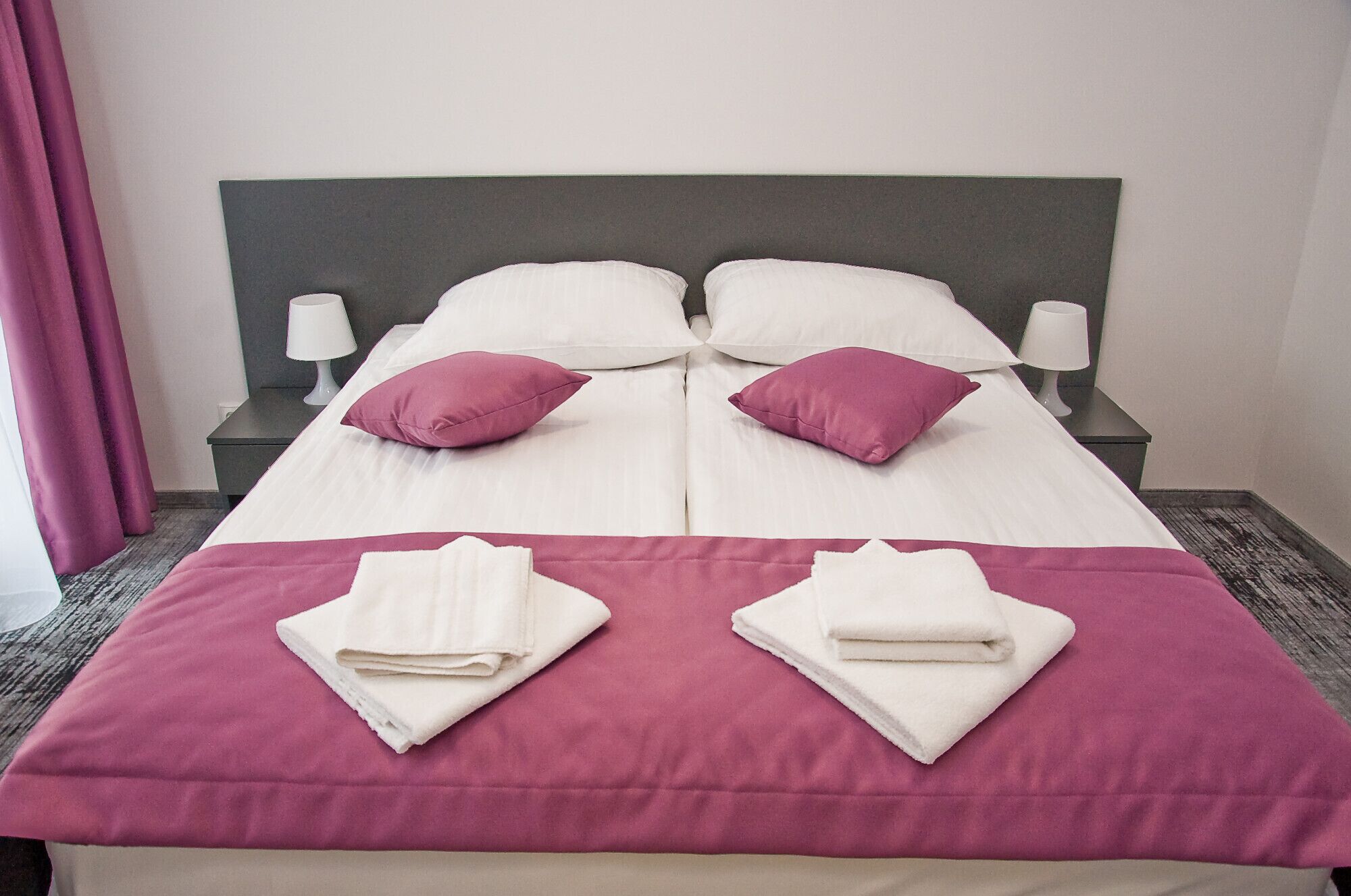 Un llit ben arreglat amb roba de llit blanca i rosa, dues tovalloles blanques doblegades sobre el llit, i dues làmpades de nit sobre taulells a joc.