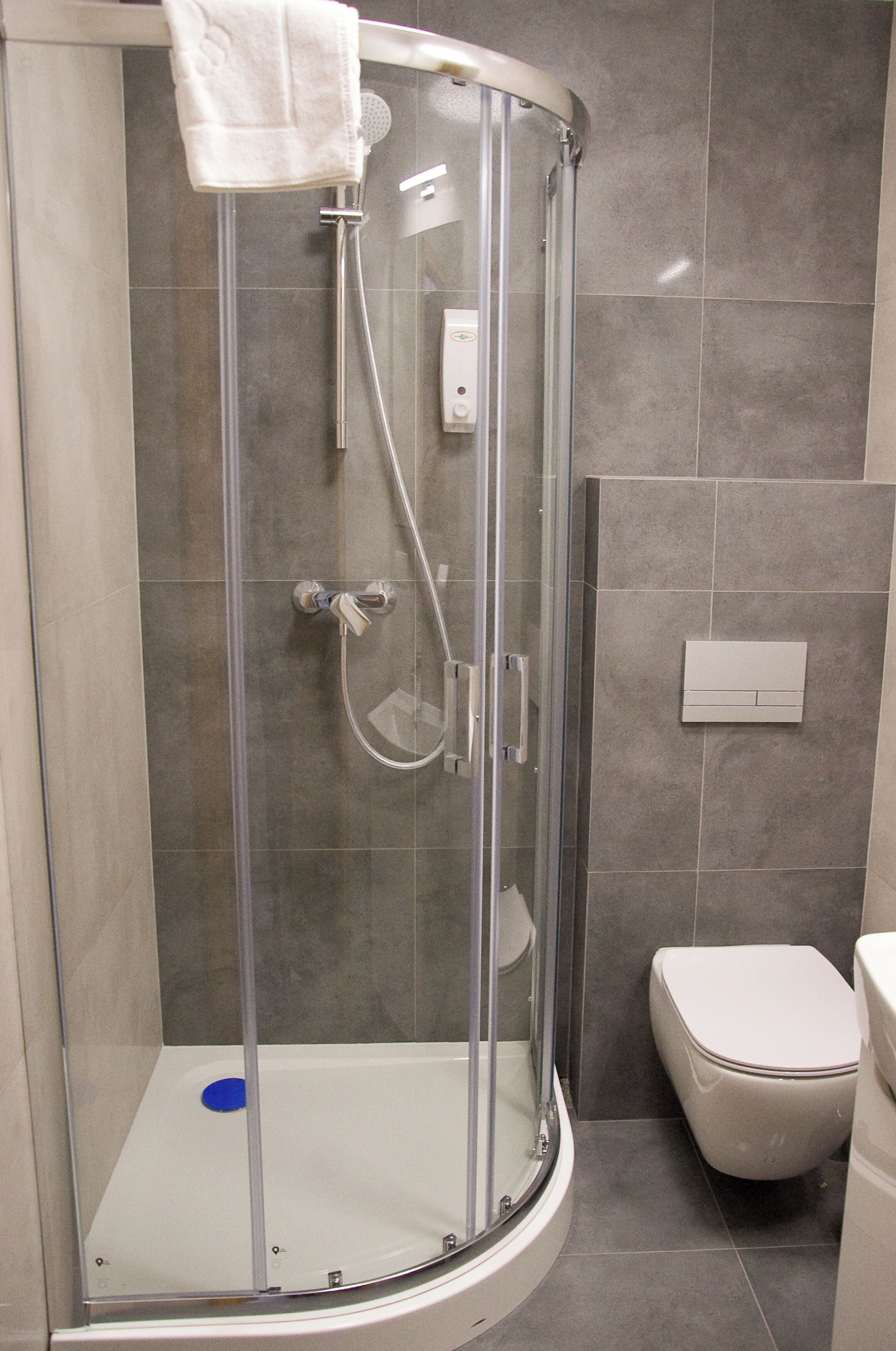 Un box de ducha con paredes de vidrio, una toalla blanca colgada en la puerta, un cabezal de ducha de mano y un panel de control en la pared.