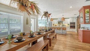 A modern kitchen with a wooden dining table set for six, a central island with bar stools, and a view into the adjacent living area.
