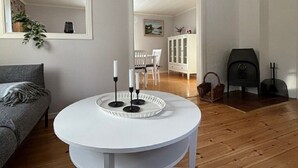 A living room with a round white table, a grey sofa, a fireplace, and a dining area in the background. - Cozy Swedish Waldhof Marie - in a secluded location - fireplace & nature (Västra Götalands län)