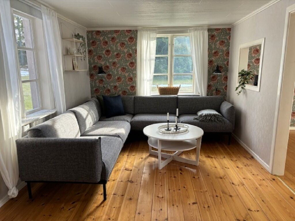 A living room with a grey sectional sofa, a white coffee table, wooden flooring, floral wallpaper, and a window with curtains.
