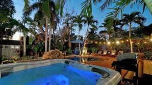 A hot tub surrounded by palm trees and a small waterfall, with lounge chairs and string lights.