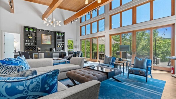 A spacious living area with a large sofa, a coffee table, and a dining area with chairs. There is a kitchen area with a bar and shelves with decorative items. The room has large windows and a view of the outdoors. - Luxurious Retreat | Private Creek View | Hot Tub (Hot Springs)
