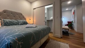Ein Schlafzimmer mit einem gepolsterten Kopfteil, einem Bett mit gemusterten Betttextilien, einer Nachttischlampe, einem begehbaren Kleiderschrank, einem roten Sessel und einer Topfpflanze.