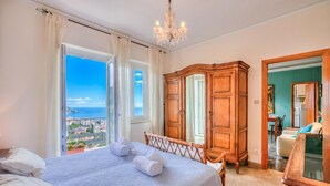 A bedroom with a bed, a wooden wardrobe, a chandelier, and a view of the sea. - Simply Romantic Alassio Sea View (Alassio)