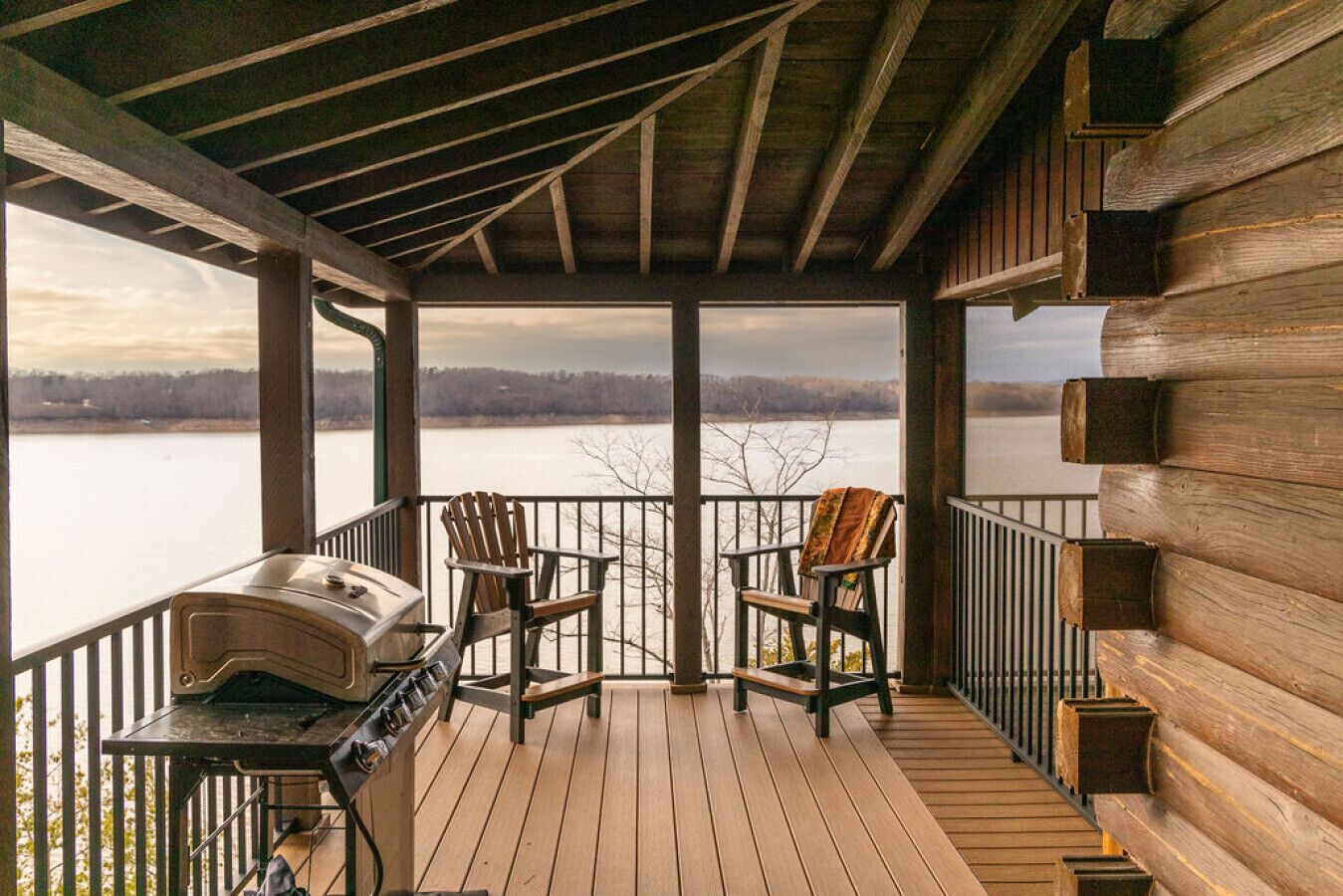 Una terraza de madera con una parrilla y dos sillas Adirondack con vistas a un lago.