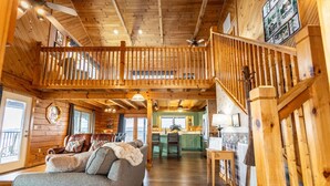 Un intérieur spacieux de chalet en bois, avec un plafond haut, un balcon, un coin salon confortable avec un canapé et une cuisine à l’arrière-plan.