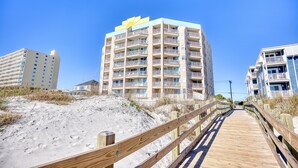 A wooden boardwalk leading to a beachfront building with multiple balconies and a yellow logo on the top.