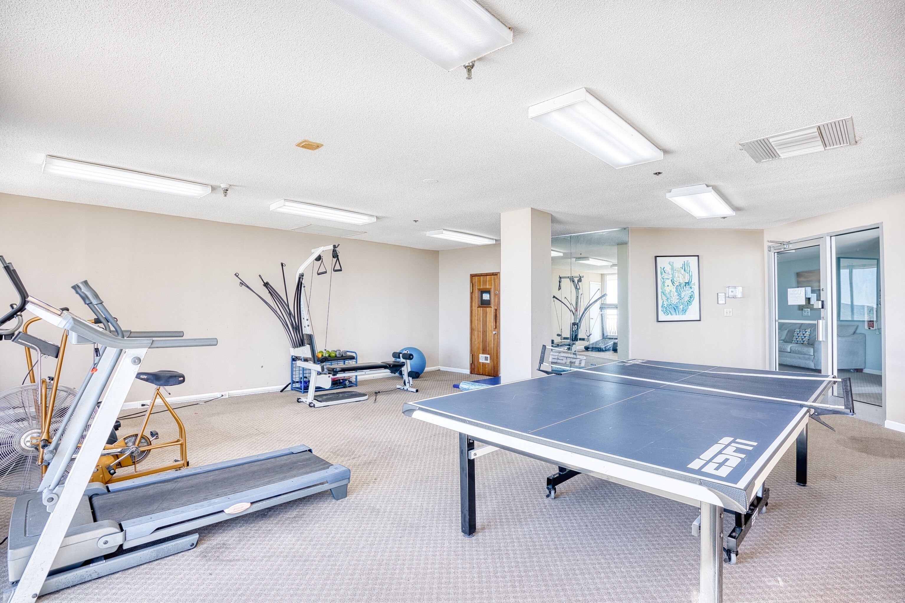 Ein Fitnessraum mit Trainingsgeräten, einem Tischtennis-Tisch und einem Spiegel.