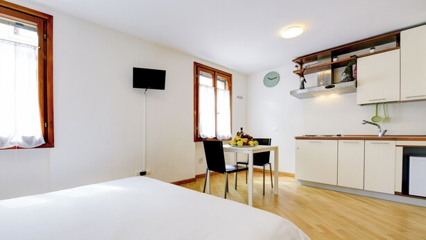 A room with a bed, a dining table with chairs, a kitchen area with cabinets, a clock, and a television.