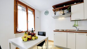 A small kitchen with a white table, a fruit bowl, a clock, and a window with curtains.