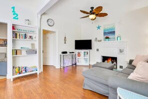 A living room with a grey sofa, wooden flooring, a bookshelf, a flat-screen TV, a fireplace, and a ceiling fan. - Spacious Home with Bikes, Near Historic Downtown (St. Augustine)