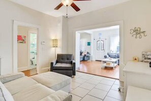 A spacious living room with a black leather chair, a white sofa, and a coffee table. There is a ceiling fan, a lamp, and a door leading to another room.