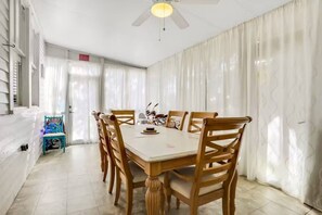 A dining area with a white table and wooden chairs, a ceiling fan, and a view of the outside through large windows with white curtains.