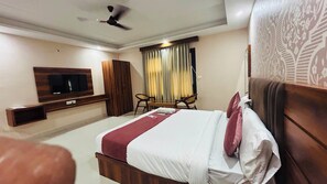 A hotel room with a bed, a TV mounted on the wall, a wardrobe, a seating area with chairs, and a ceiling fan. - Hotel Ram Janki Palace (Pindra)