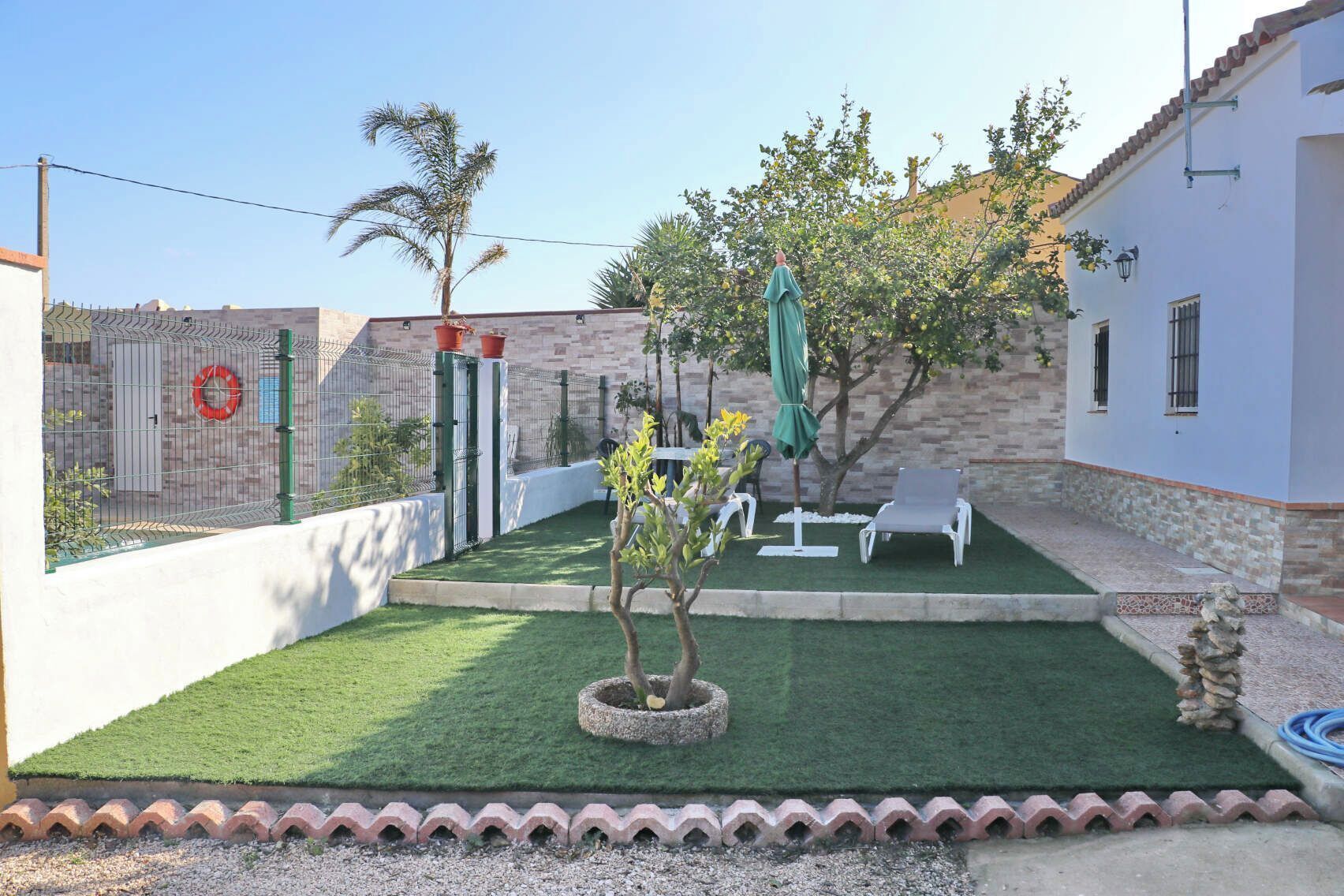 Un patio trasero cercado con césped artificial, un árbol pequeño y mobiliario exterior.