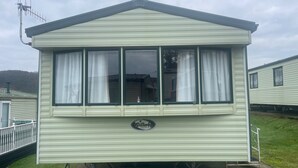 A single-story mobile home with a peaked roof and a front porch. It has a small porch with a railing and a window with curtains. The home is situated on a grassy area with a paved path leading to it. - Seafront Cozy Static Caravan With Sea Vievs (Aberystwyth)
