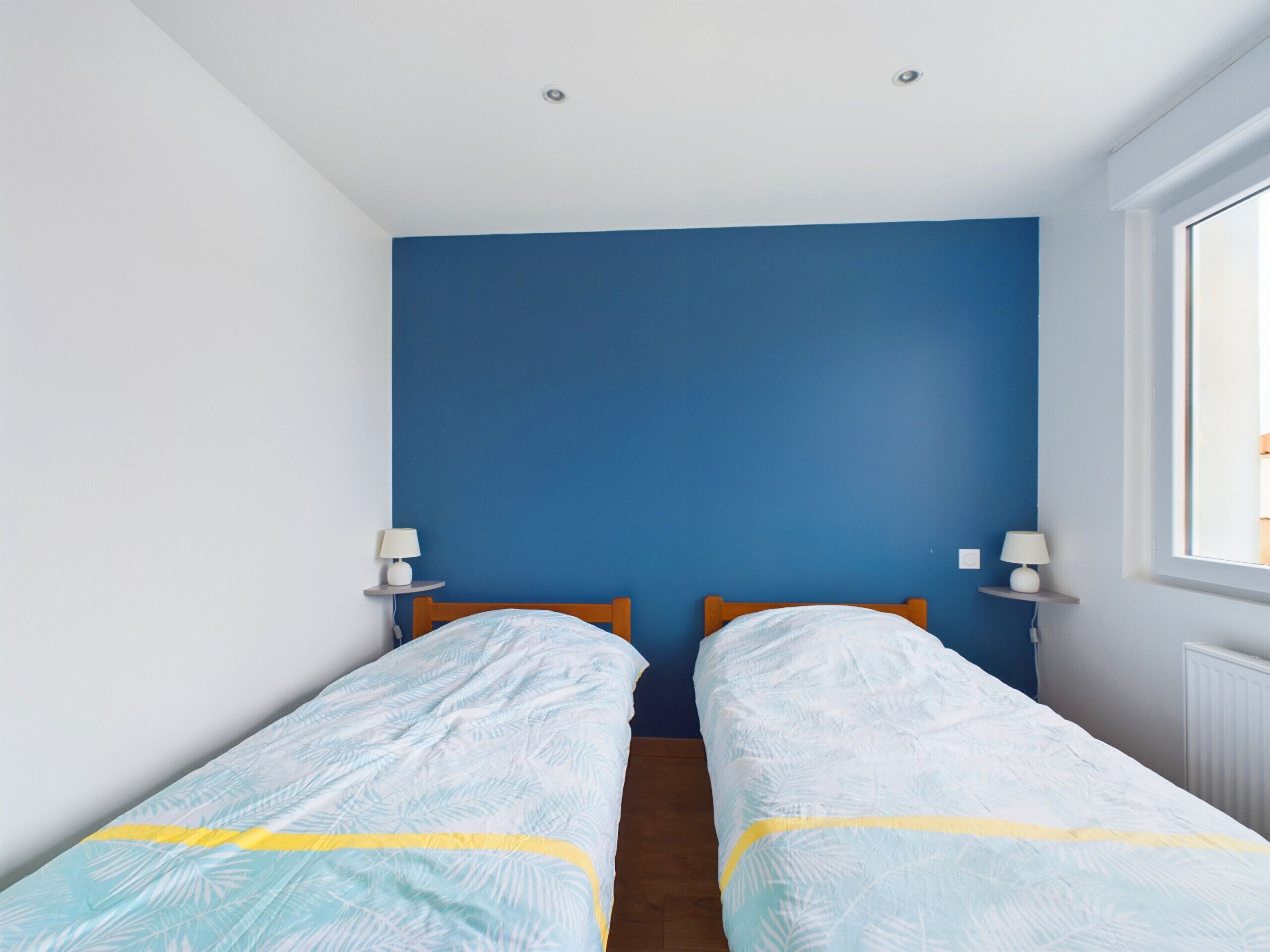 Une chambre avec deux lits simples, un mur d  ’accent bleu et une fenêtre laissant entrer la lumière naturelle.