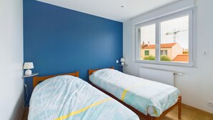 A bedroom with two beds, a window with a view of buildings, and a blue wall. - House with garden, beach 15 min walk - 6 guests (Les Sables-d´Olonne)