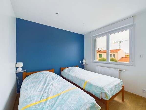 A bedroom with two beds, a window with a view of buildings, and a blue wall.