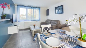 A modern living room with a sofa, a dining table set for two, a TV mounted on the wall, and a window with blue curtains. - House with garden, beach 15 min walk - 6 guests (Les Sables-d´Olonne)