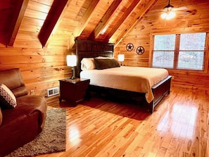 A cozy wooden cabin bedroom with a bed, nightstands, a chair, and a window with blinds.