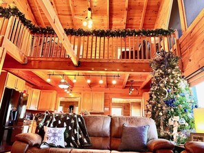A cozy cabin interior with a wooden ceiling, a dining area, a fireplace, and a Christmas tree.