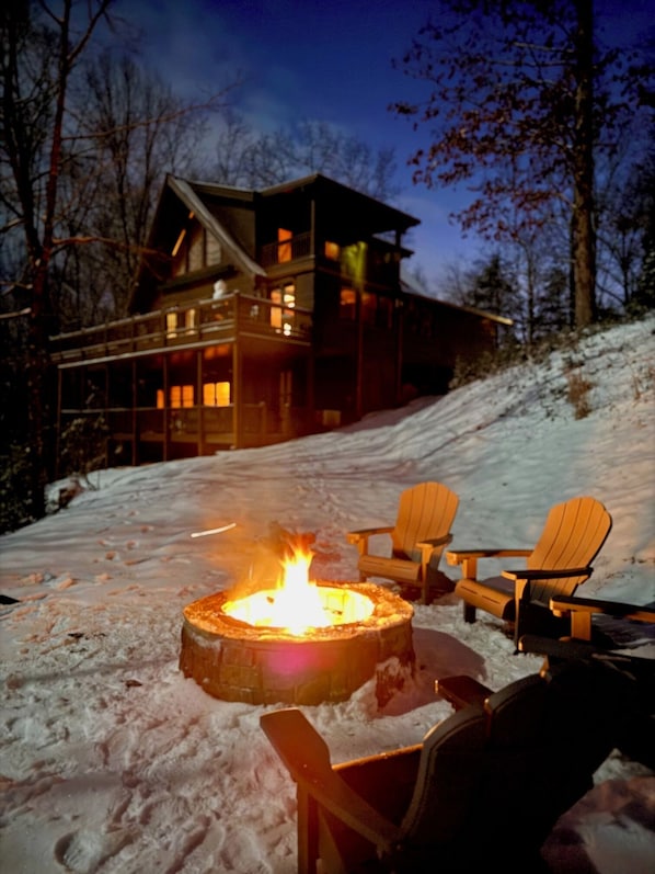A snow-covered outdoor area with a fire pit and Adirondack chairs.
