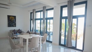 A spacious living area with a dining table and chairs, a sofa, and large windows with curtains. - Aika House - A haven of peace in the heart of Michamvi  (Michamvi Kae.)