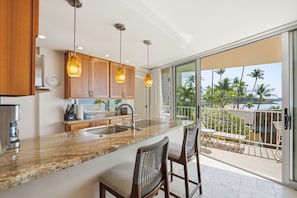 A kitchen with a large island, wooden cabinets, and pendant lighting. There is a view of a balcony with outdoor furniture.