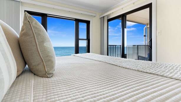 A bedroom with a large bed, a view of the ocean through open windows, and a balcony with a railing. - Oceanfront Condo with Stunning Views, Balcony, & Steps to the Beach! (Ocean City)