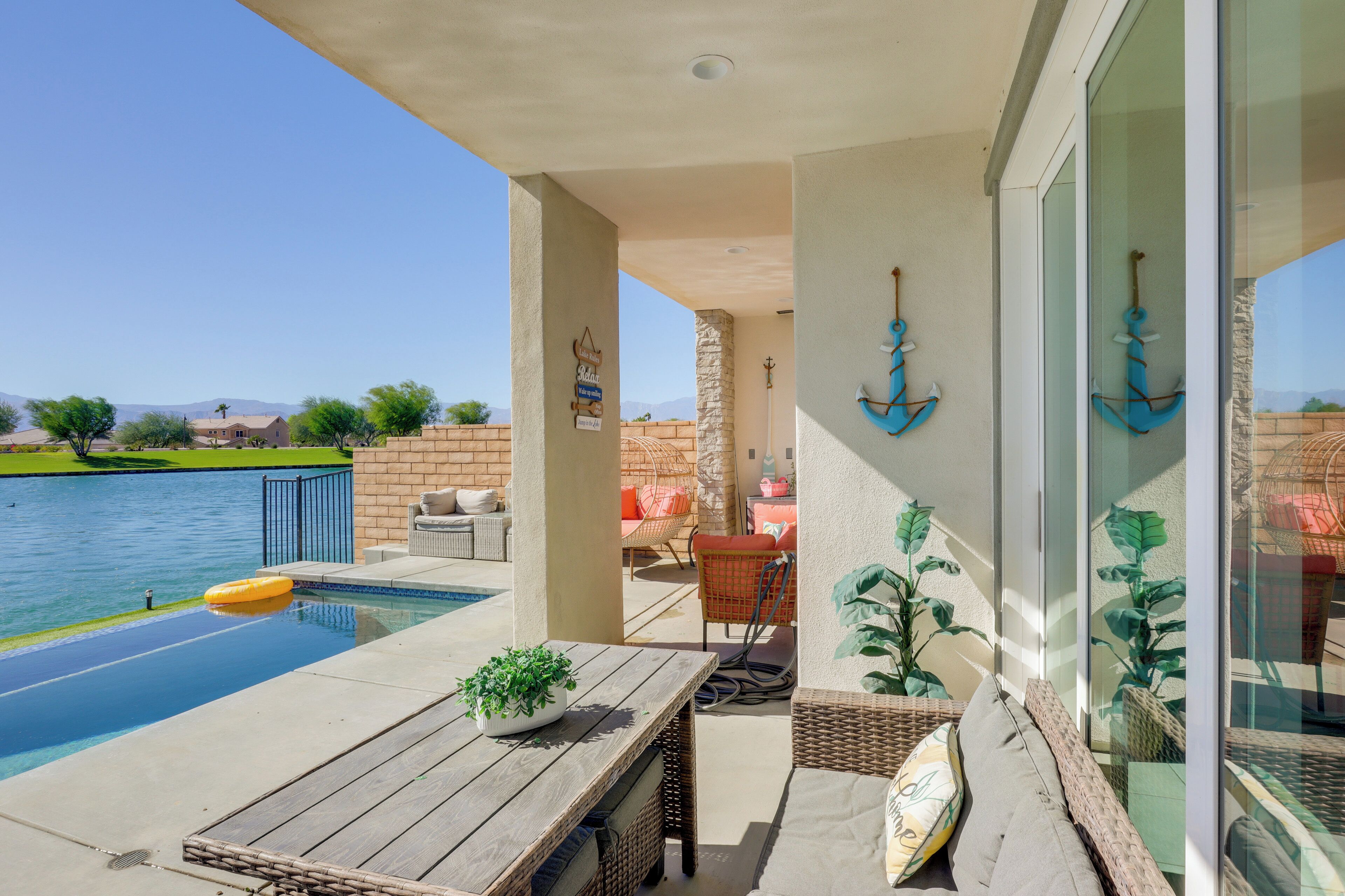 Zona junto a la piscina con mesa de madera, mobiliario de mimbre y vista a un lago.
