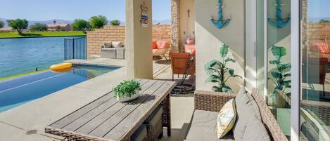 Zona junto a la piscina con mesa de madera, mobiliario de mimbre y vista a un lago.