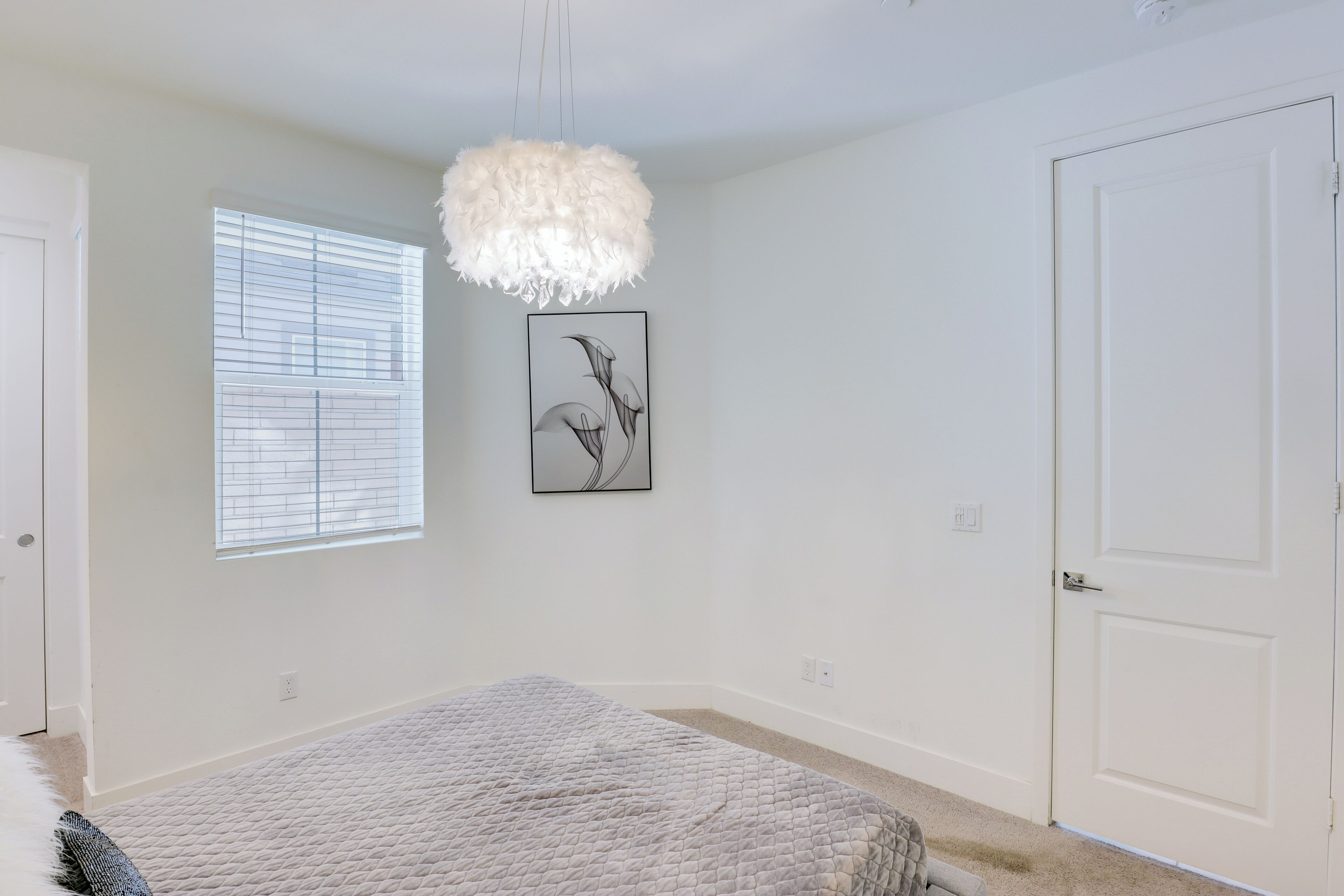 Un dormitorio con cama, una puerta blanca, ventana con persianas, una obra de arte en la pared y una lámpara colgante blanca y mullida.