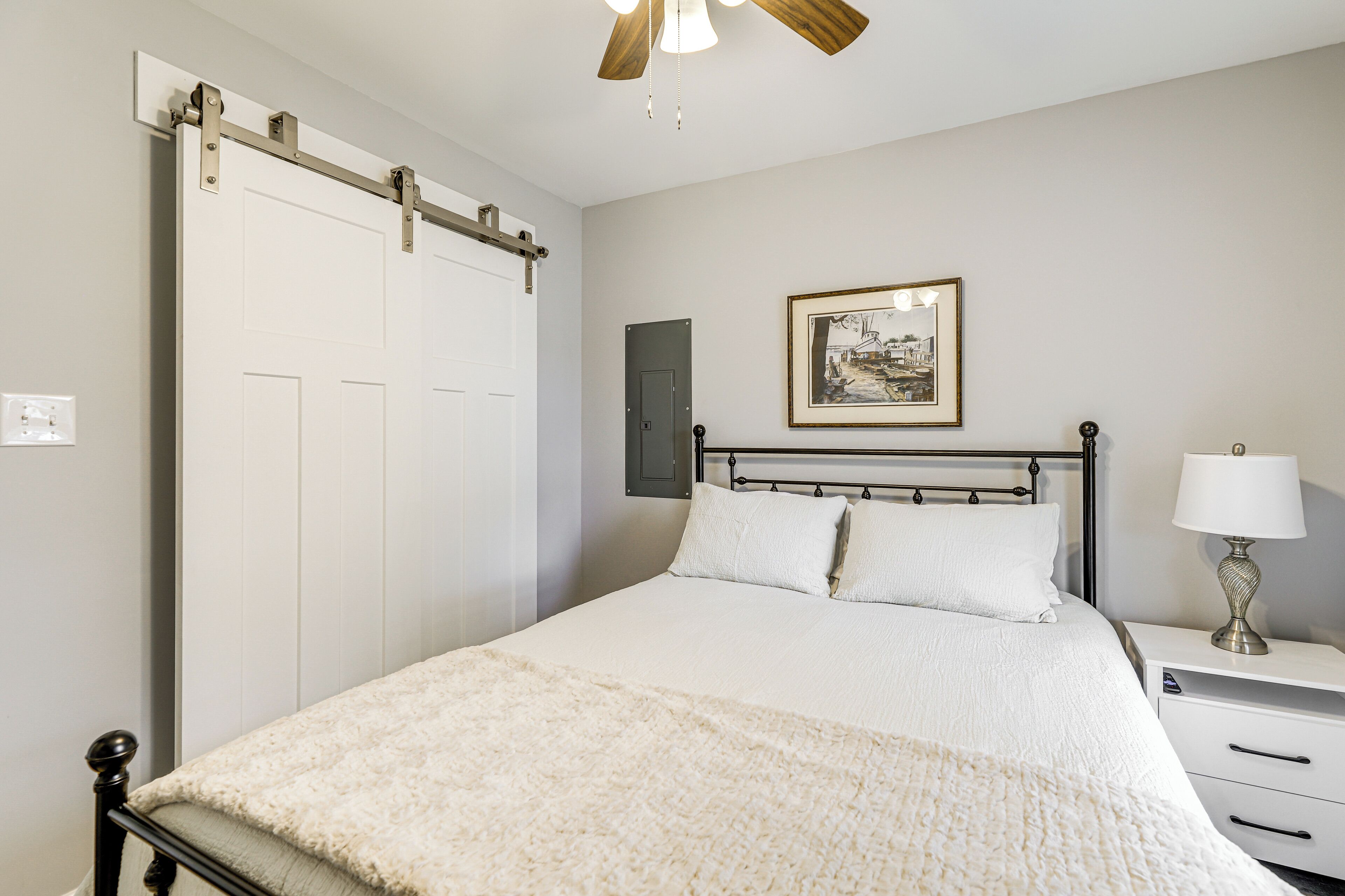 A bedroom with a bed, a nightstand with a lamp, a framed picture on the wall, and a sliding door.