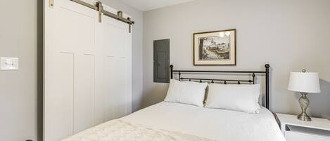 A bedroom with a bed, a nightstand with a lamp, a framed picture on the wall, and a sliding door.