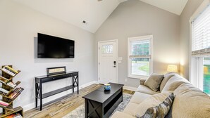 Un salon avec une console noire, un canapé beige, un téléviseur à écran plat fixé au mur et une fenêtre avec des stores.