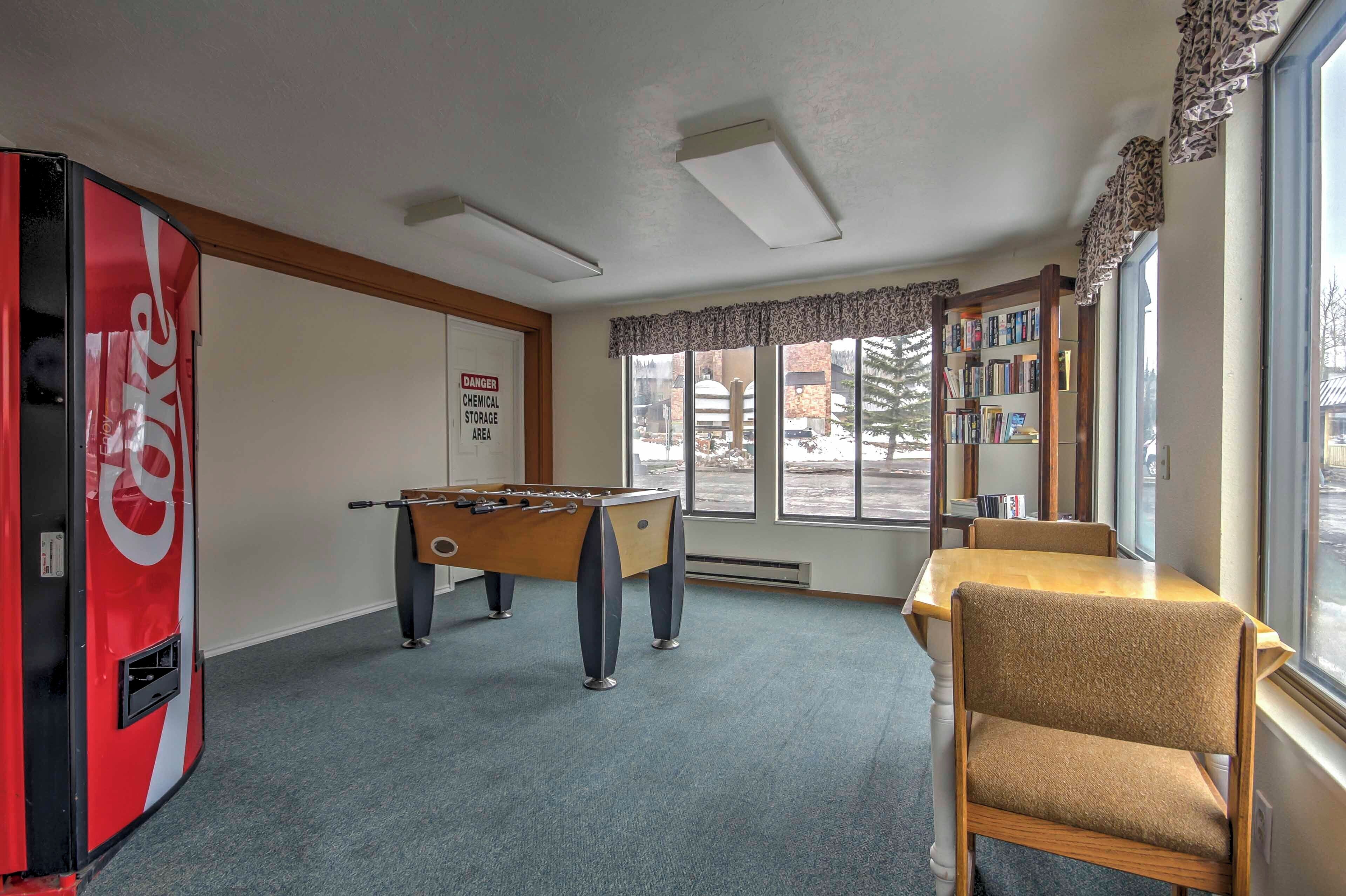 A room with a foosball table, a Coca-Cola refrigerator, a bookshelf, and a window with a view of the outside.