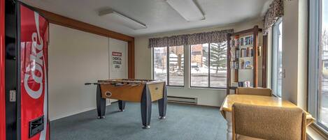 A room with a foosball table, a Coca-Cola refrigerator, a bookshelf, and a window with a view of the outside.