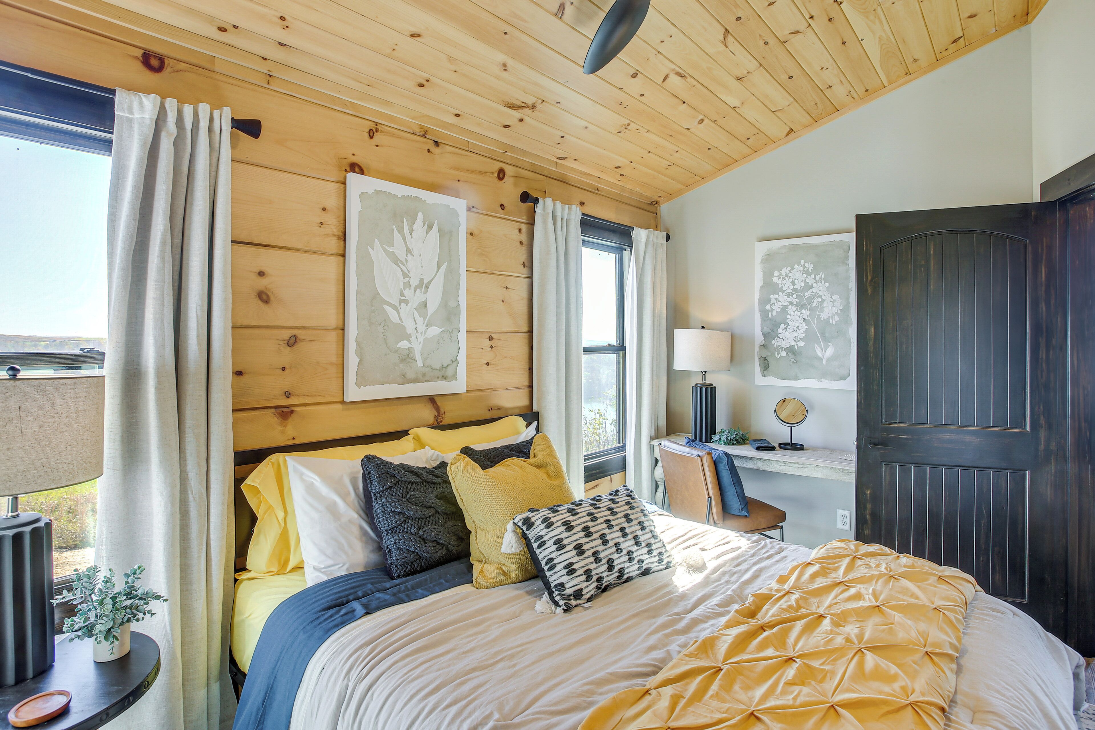 A bedroom with a wooden ceiling, a bed with yellow and white bedding, a bedside table with a lamp, and a window with curtains.
