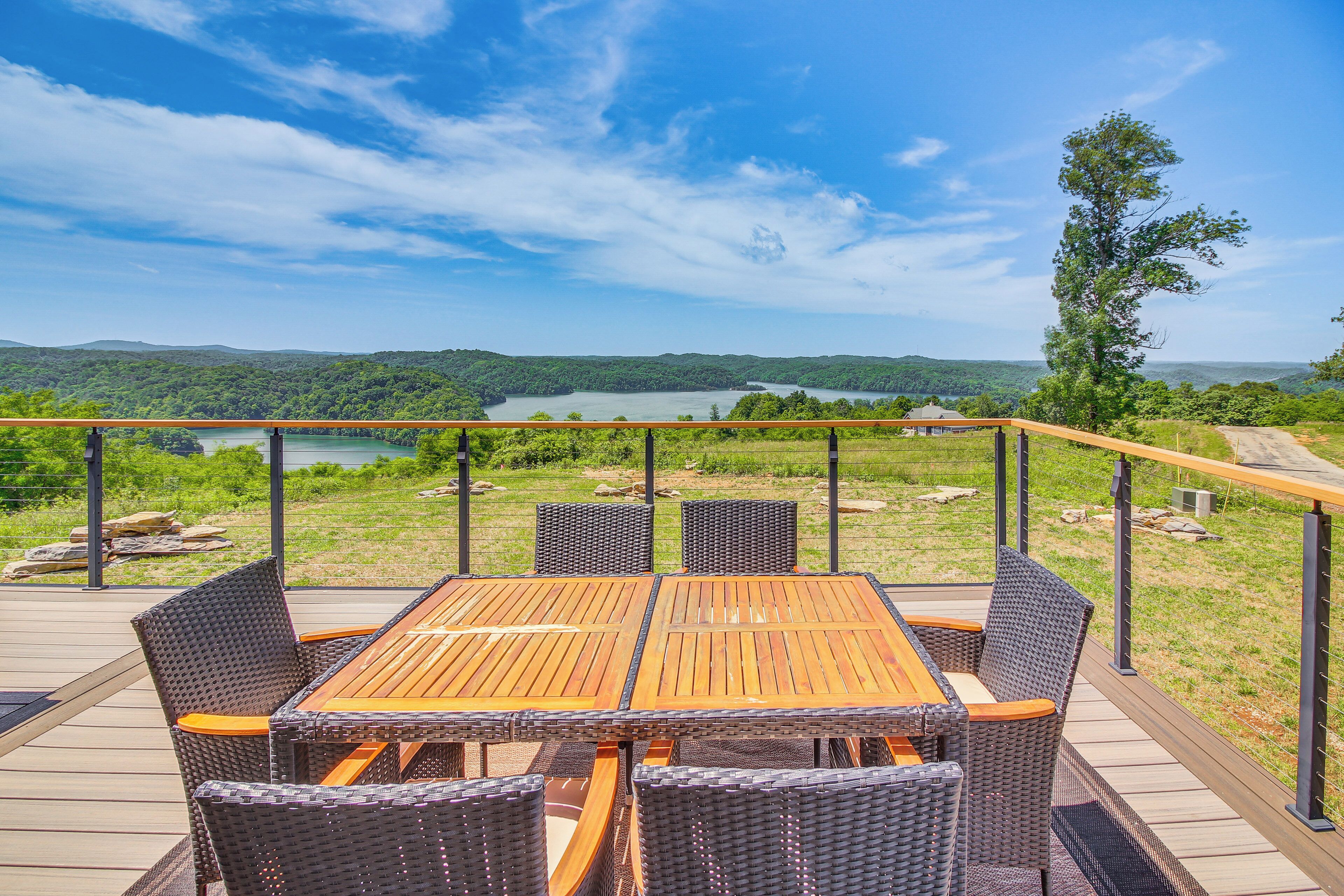Een houten terras met tafel en stoelen met uitzicht op een meer en beboste heuvels.