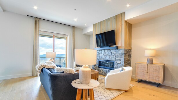 A modern living room with a stone fireplace, a flat-screen TV mounted above it, and a large window with curtains. - Ski-in/out Townhome w/ Views at Okemo Resort! (Ludlow)