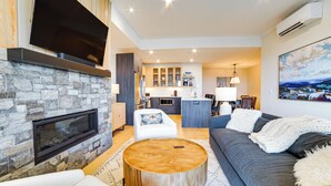A modern living room with a stone fireplace, a flat-screen TV, a grey sofa, and a wooden coffee table. - Ski-in/out Townhome w/ Views at Okemo Resort! (Ludlow)
