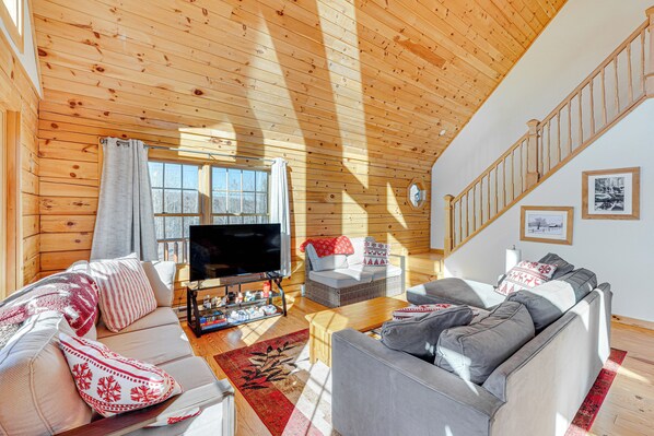A cozy living room with a wooden ceiling, a flat-screen TV, two sofas with patterned cushions, a coffee table, and a staircase leading to another level. - Cabin 4 Mi to Belleayre Mountain: Ski, Hike & Fish (Fleischmanns)