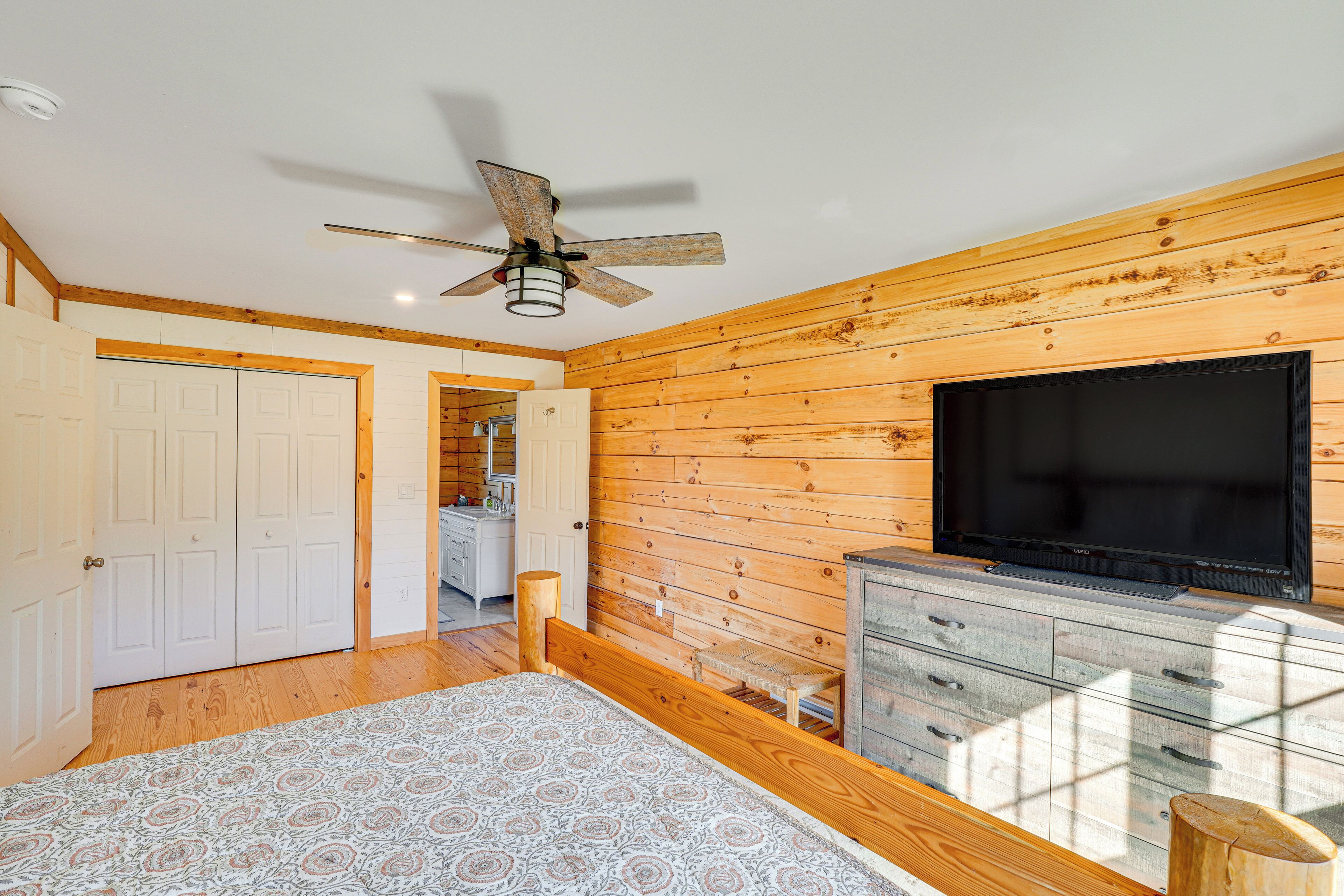 A bedroom with a bed, a ceiling fan, a TV, and wooden walls.